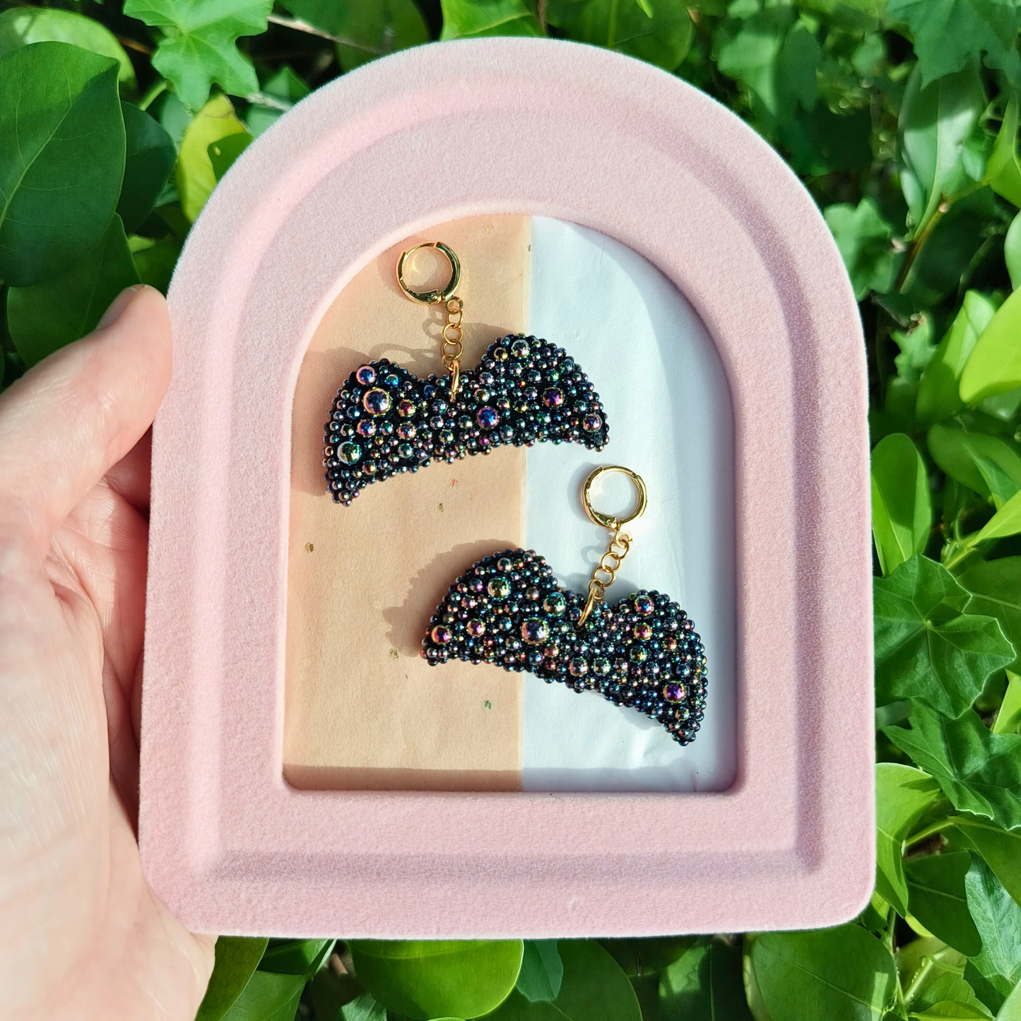 A pair of black bat-shaped earrings with onyx stones, displayed in a pink and beige case, against background of bushes
