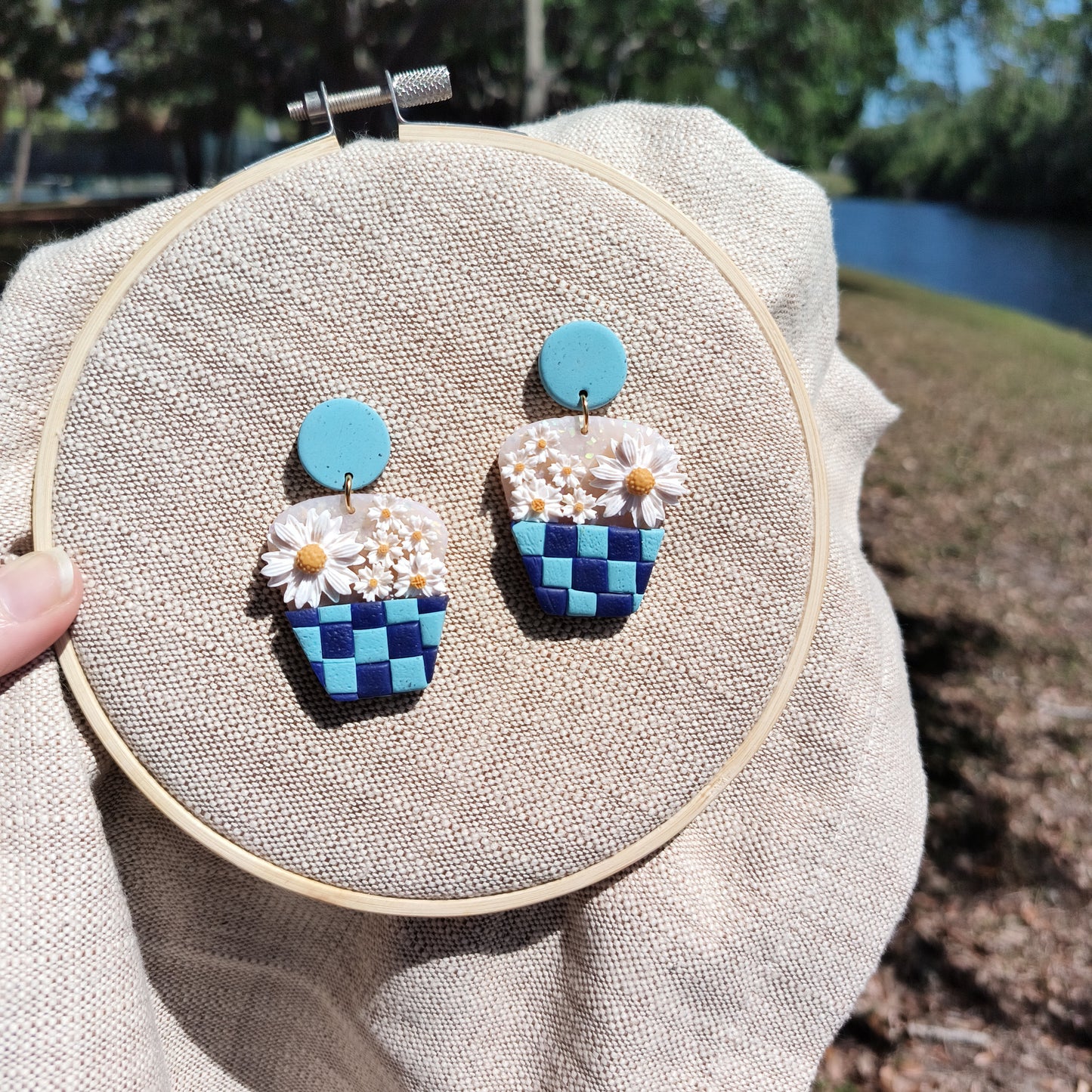 polymer clay earrings of checkerboard baskets filled with varying sizes of daisies. displayed on an embroidery hoop, with a pond in the background.