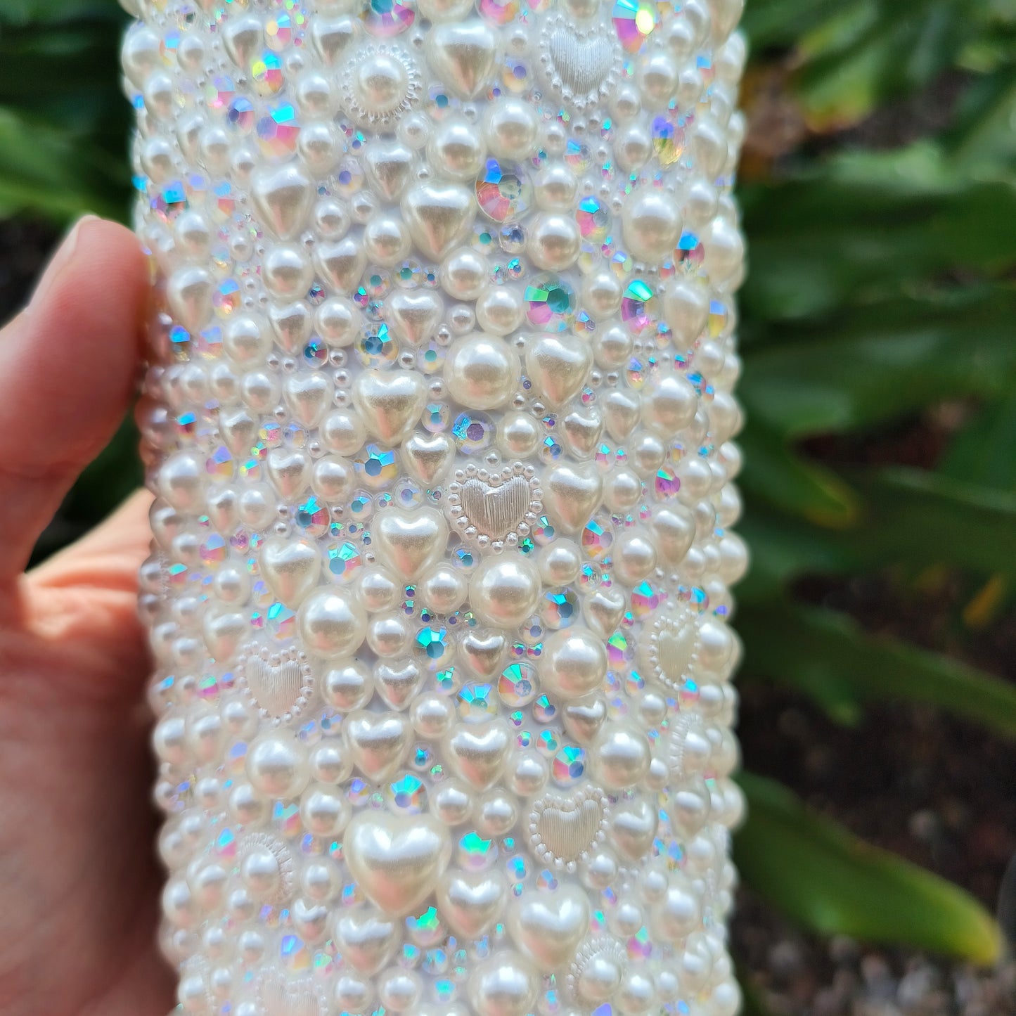 photo of the small details of bedazzled white tumbler. Shows pearl hearts of all sizes, tiny pearls, sparkly rhinestones of varying sizes. against a backdrop of philodendron.