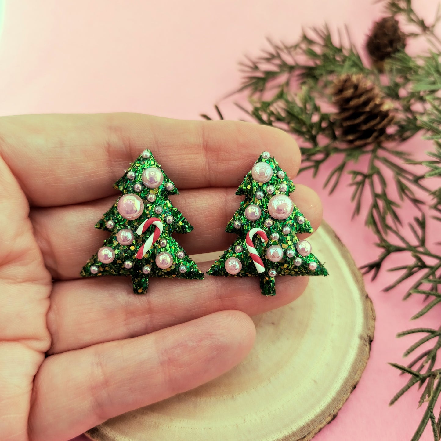 Green Christmas tree earrings with candy cane and pearl details held in a hand against a pink background with greenery.