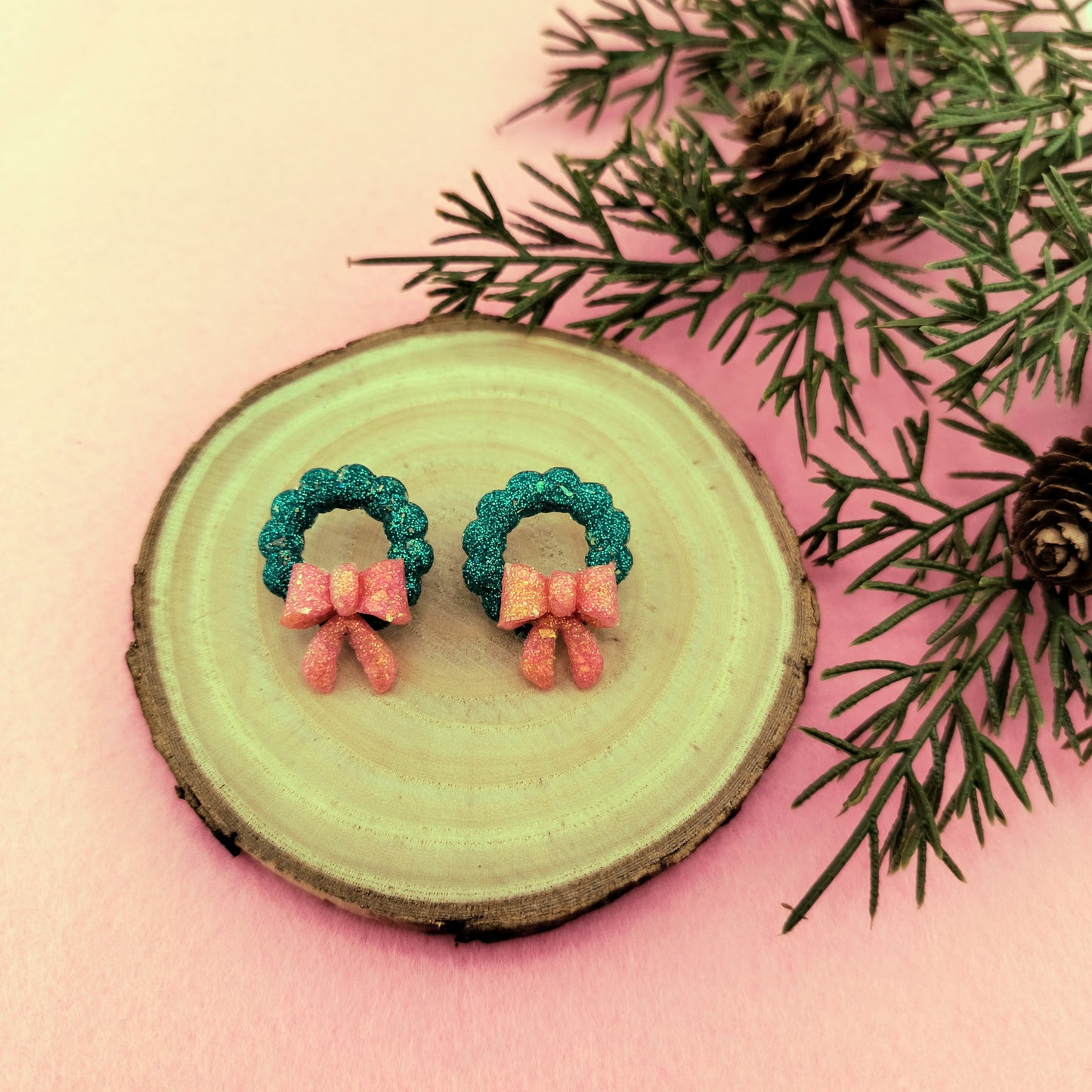 Pair of Christmas wreath earrings on a wooden slice with pine branches.