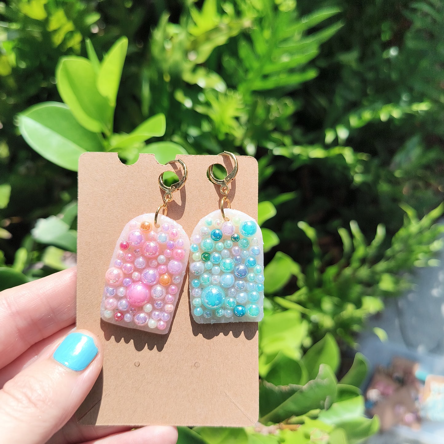 Dangle earrings covered in multi colored flat back pearls, against a backdrop of green foliage.