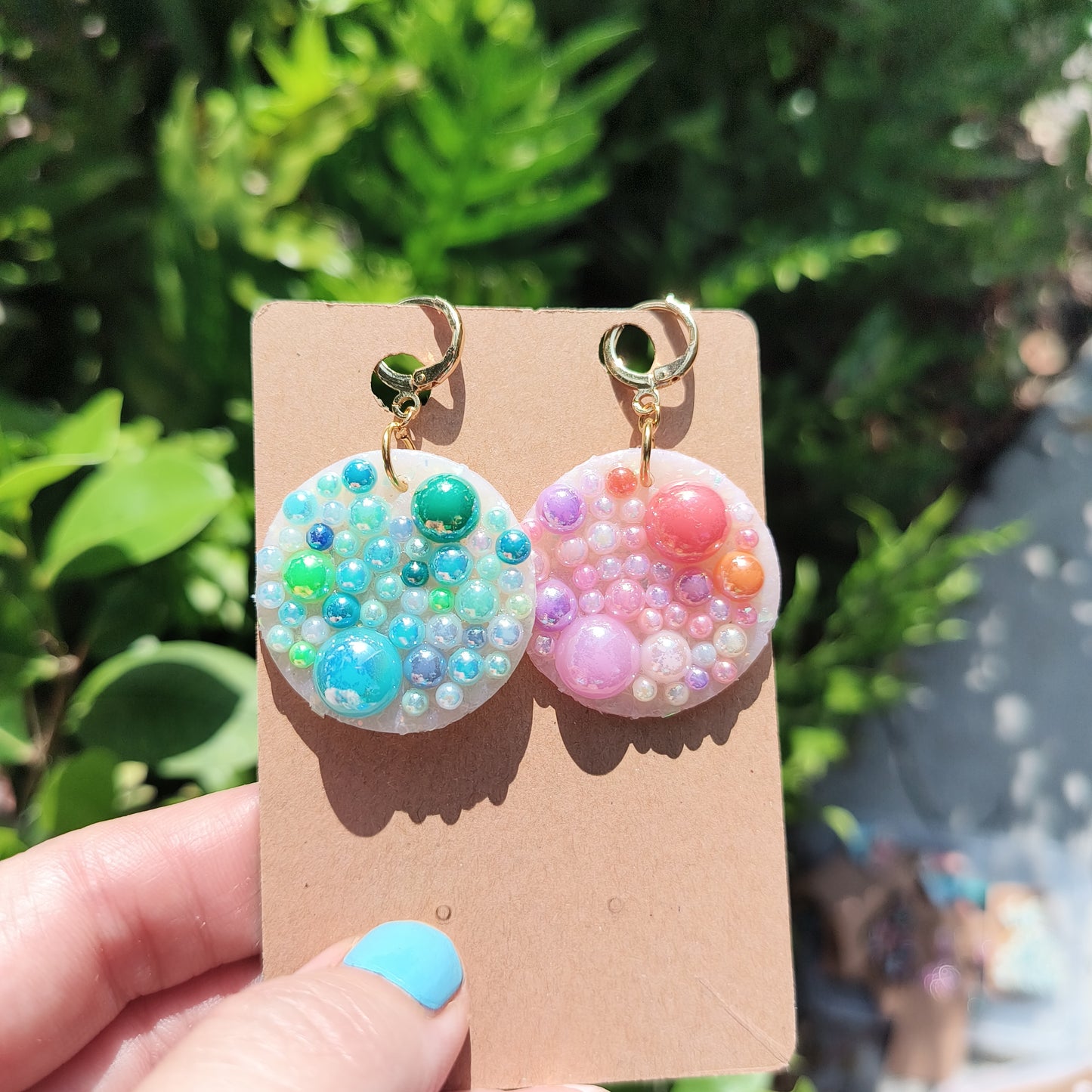 Circle dangle earrings covered in rainbow toned pearls, against a backdrop of green foliage.