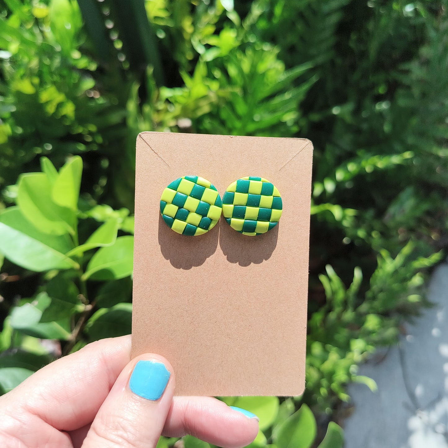 Green checkerboard polymer clay stud earrings, against a backdrop of green foliage.
