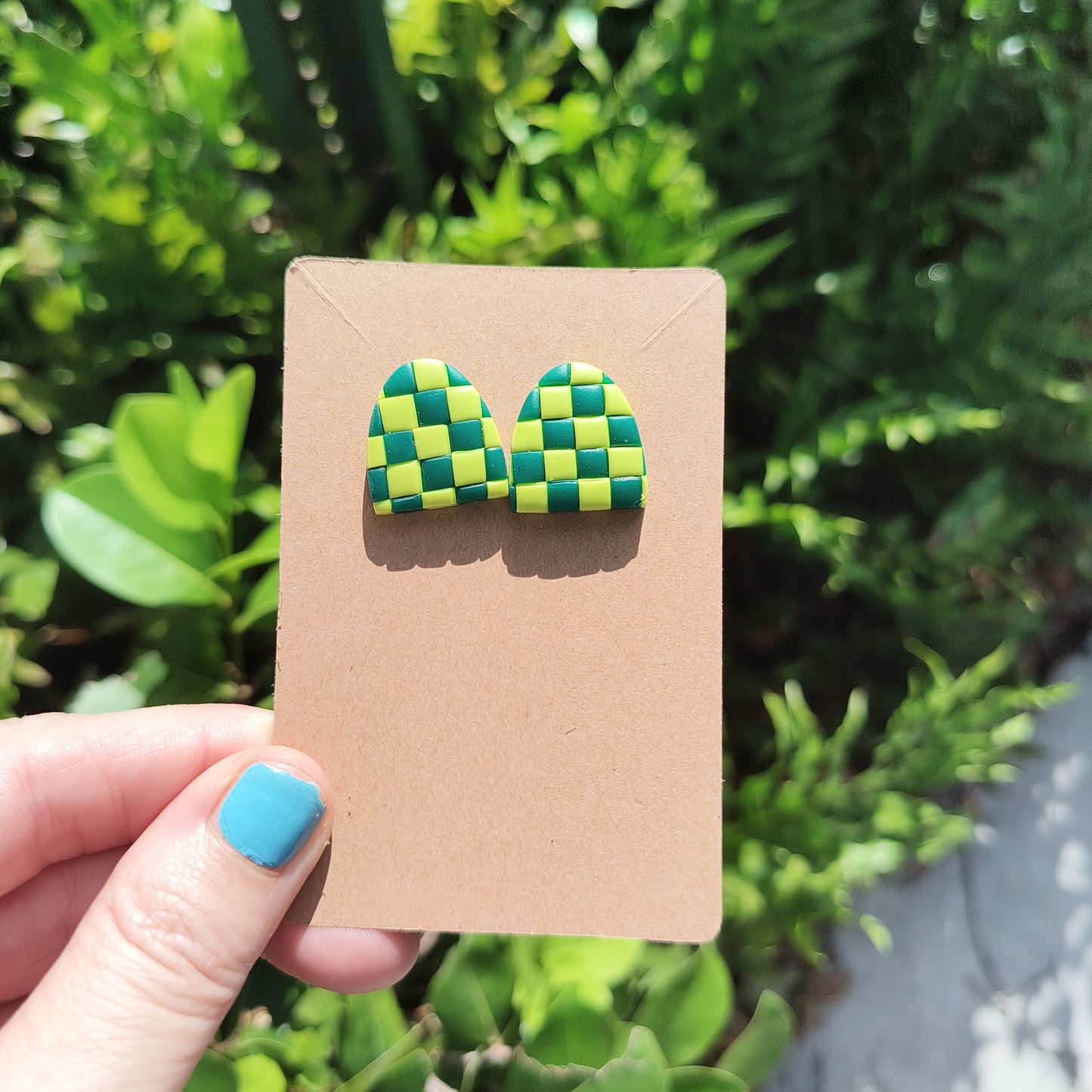 Green checkerboard polymer clay stud earrings, against a backdrop of green foliage.
