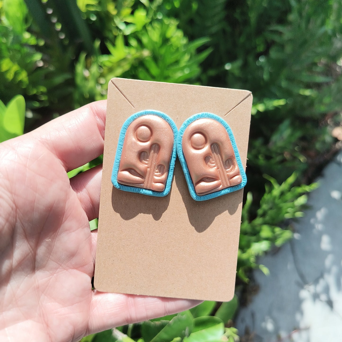 Rose gold polymer clay earrings with a desert scene stamped on it, against a backdrop of green foliage.