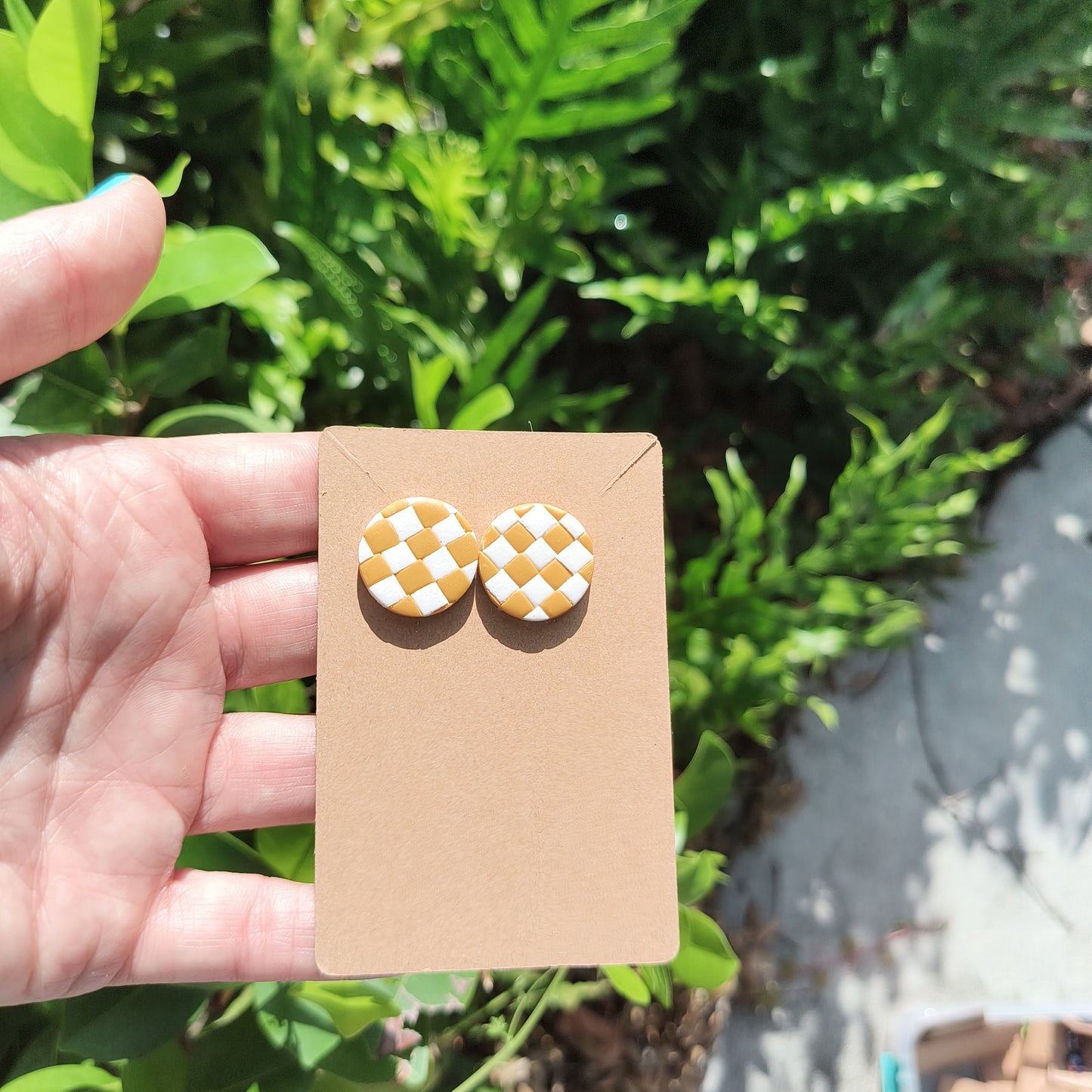White and mustard checkerboard polymer clay stud earrings against green foliage.