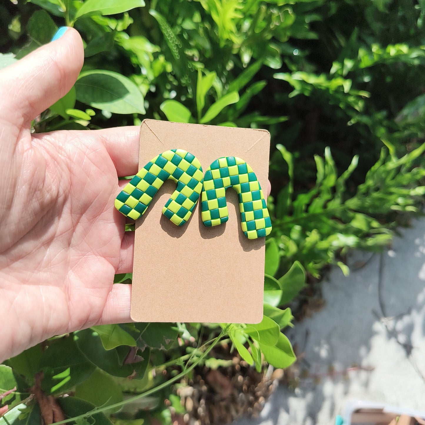 Green checkerboard polymer clay stud earrings, against a backdrop of green foliage.