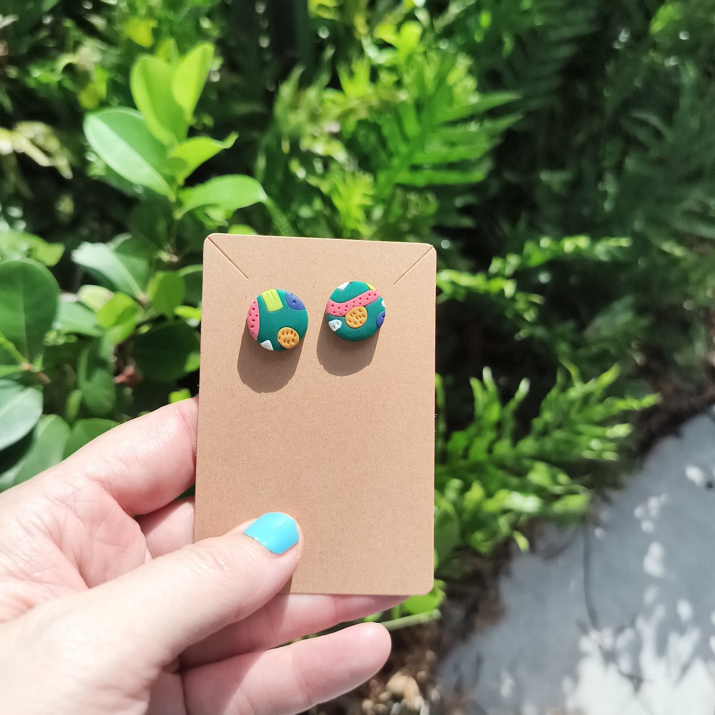 Multi colored, textured circle polymer clay stud earrings, against a backdrop of green foliage.
