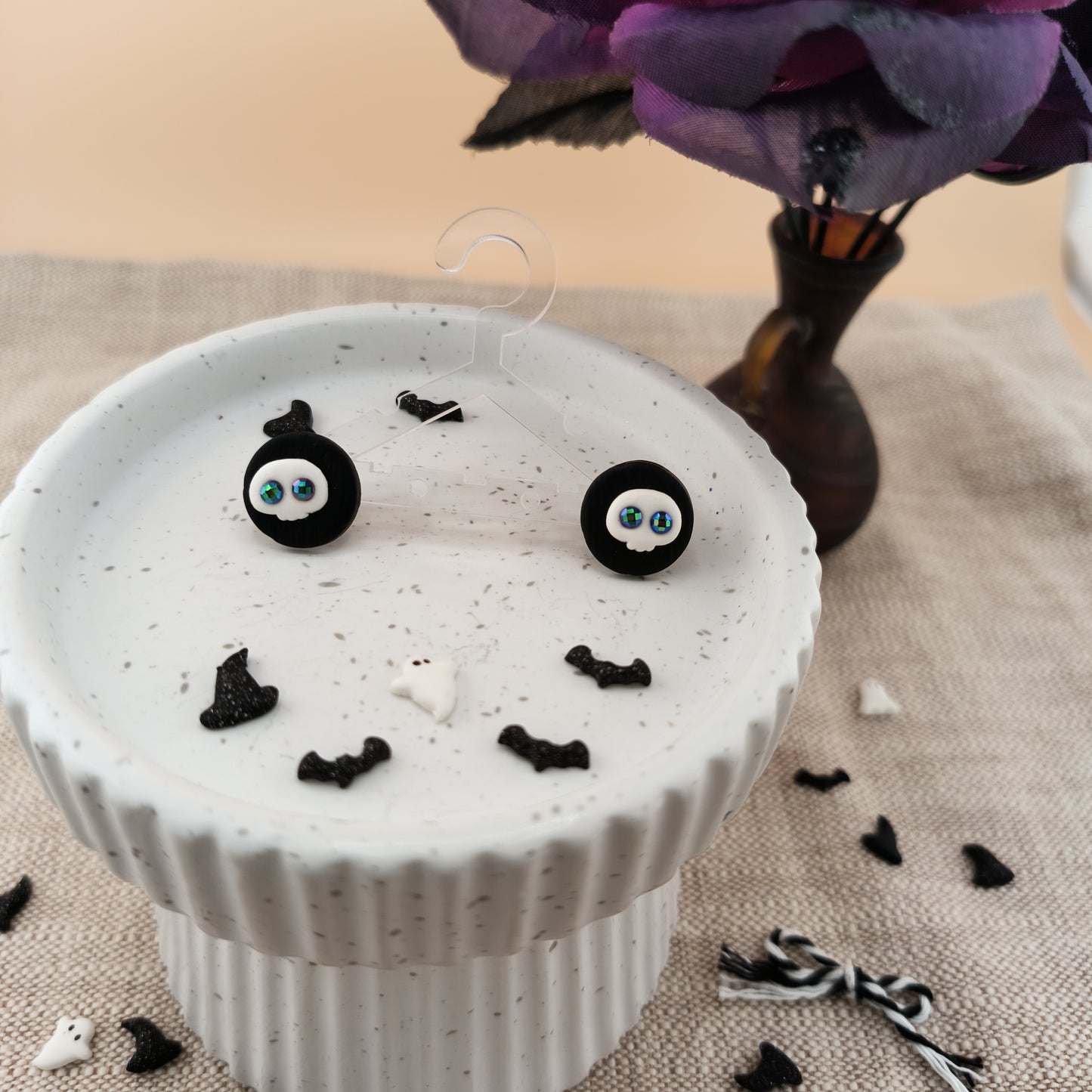 White pedestal with black bat-shaped decorations on a textured surface with a blurred flower vase in the background, featuring a pair of skull stud earrings.