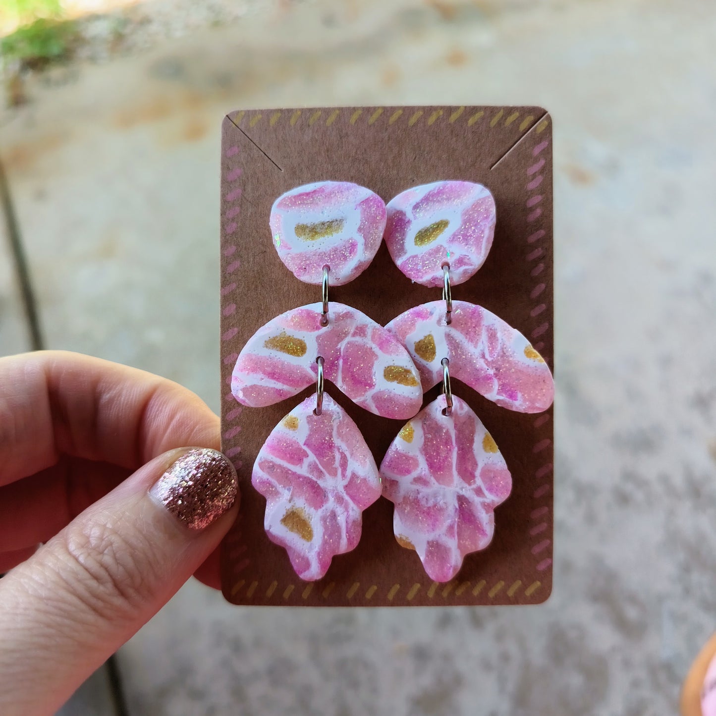 Pink butterfly-shaped earrings on a brown card held by a hand with glittery nail polish.