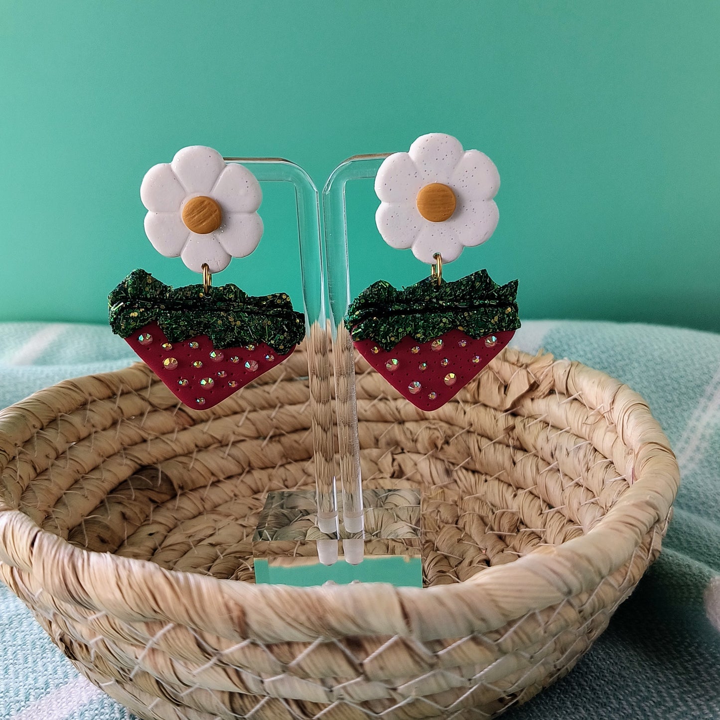 Strawberry and flower earrings on clear stands with a woven basket and teal background