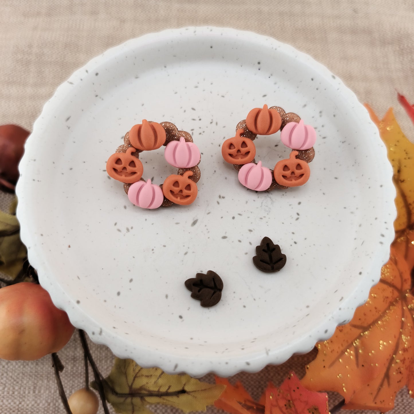 Pair of Halloween-themed earrings on a white textured surface with autumn leaves in the background.