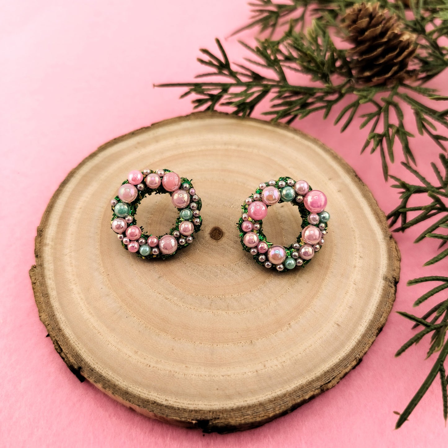 Pair of christmas wreath earrings with pink and green beads on a wooden slice with a pink background and greenery.
