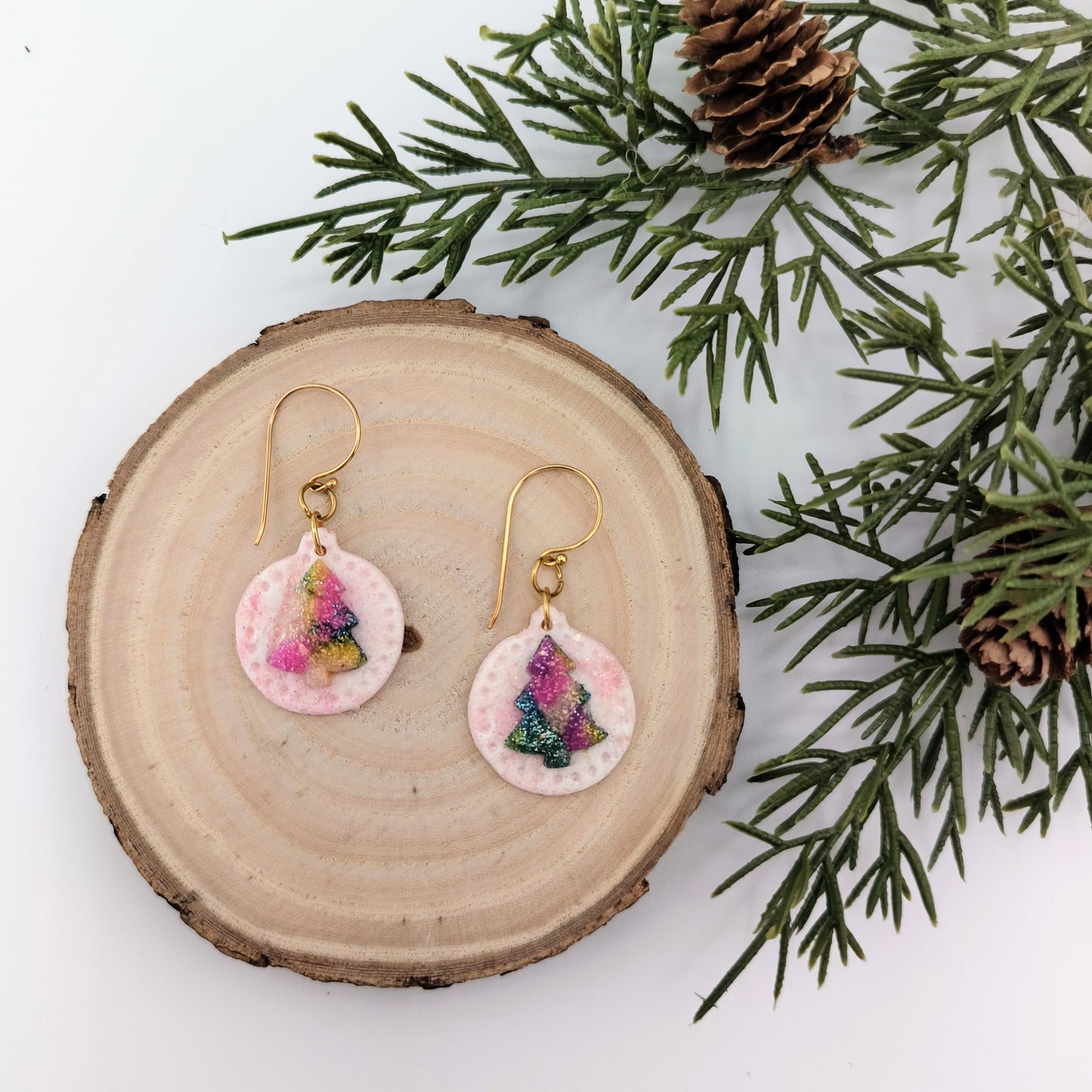 Pair of ornament earrings with colorful tree design on a wooden surface with greenery