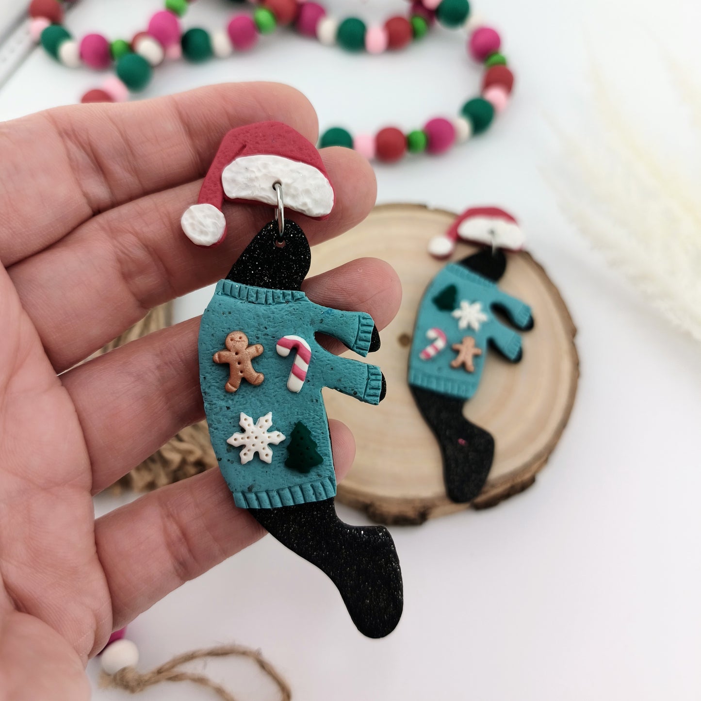 Hand holding a manatee earring wearing a blue christmas sweater and santa hat, surrounded by colorful bead garland.