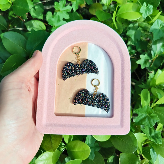 A pair of black bat-shaped earrings with onyx stones, displayed in a pink and beige case, against background of bushes