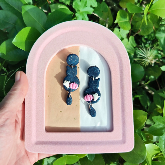 Handmade clay earrings with a cat design, with a tiny pumpkin and tiny ghost, displayed on a pink and beige stand.
