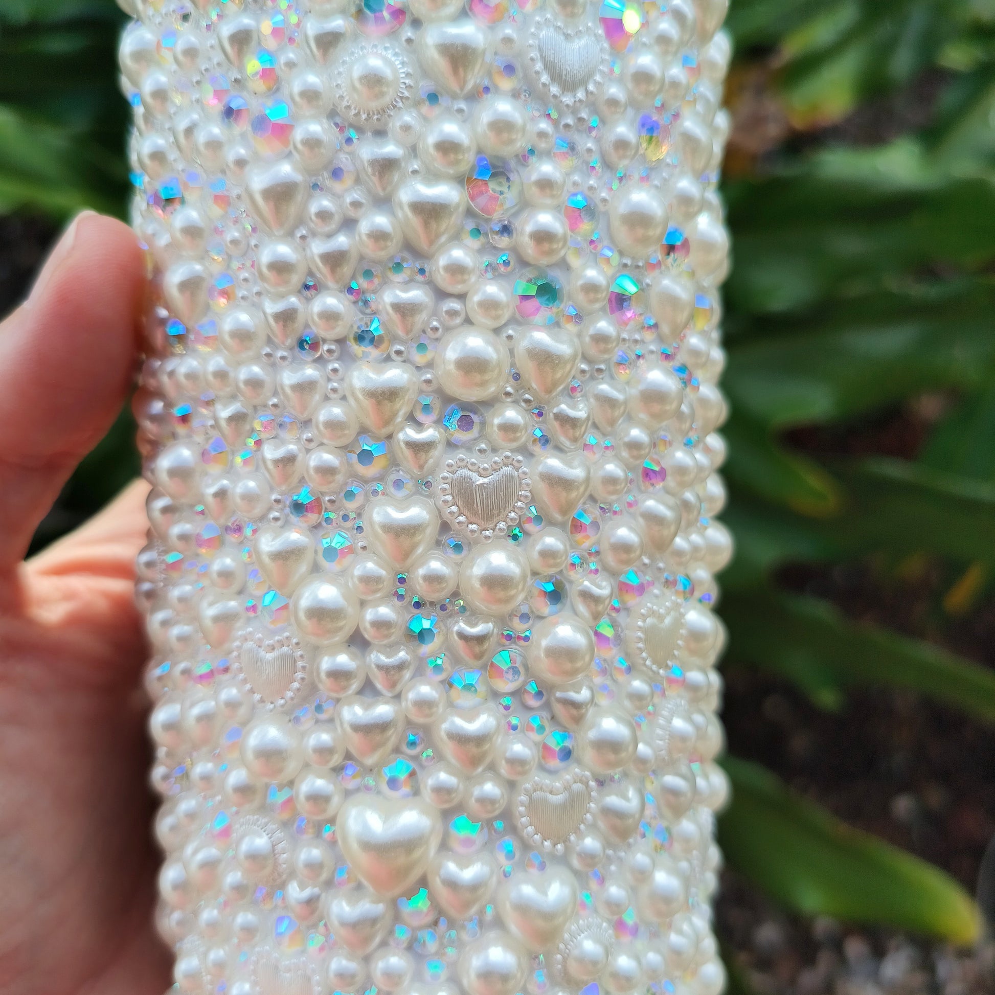 photo of the small details of bedazzled white tumbler. Shows pearl hearts of all sizes, tiny pearls, sparkly rhinestones of varying sizes. against a backdrop of philodendron. 