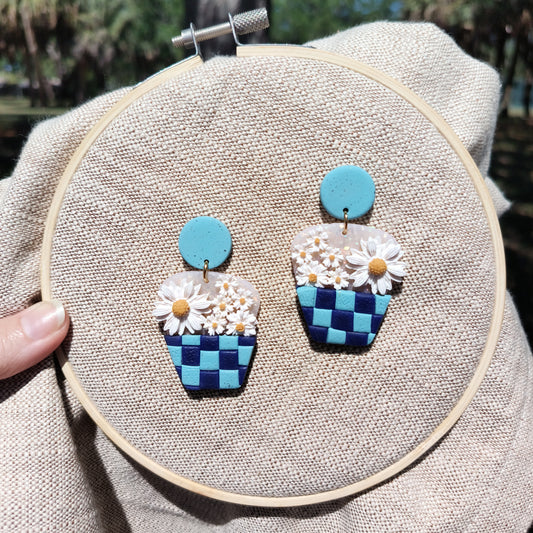 Handmade polymer clay earrings designed as a basket with daisies, displayed on a beige round base, with a blue and white color scheme.