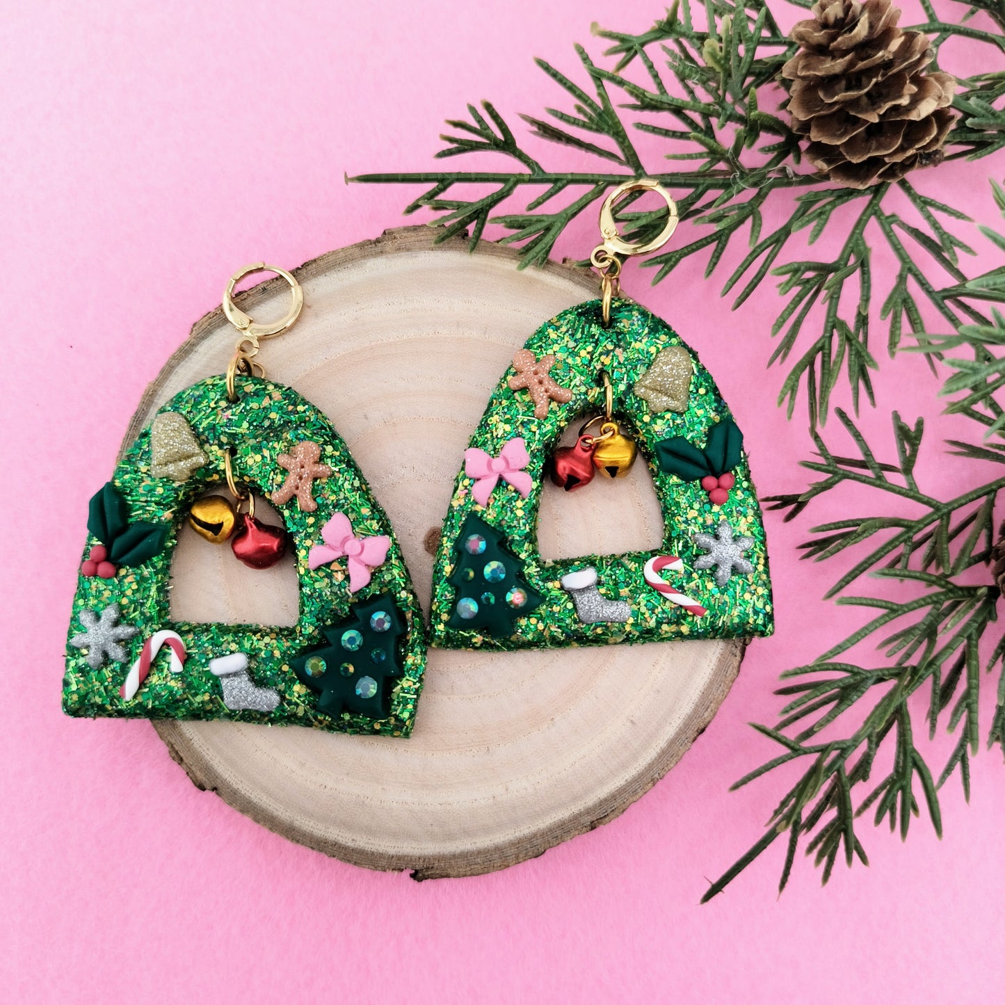 Decorative Christmas earrings with green glittery design on a pink background with pine branches.