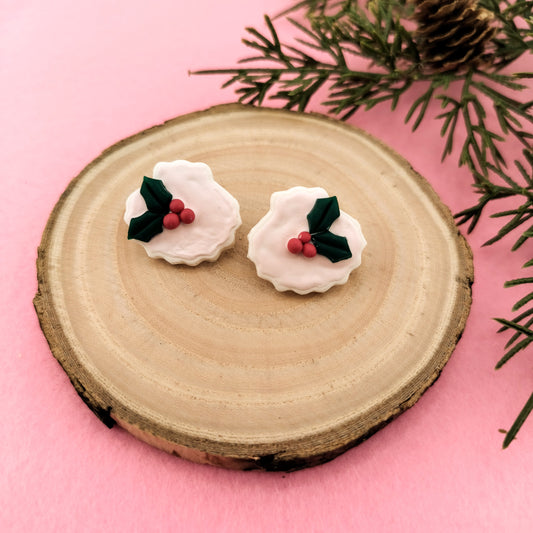 Pair of earrings resembling shells with holly leaves and berries on a wooden disc with a pink background.