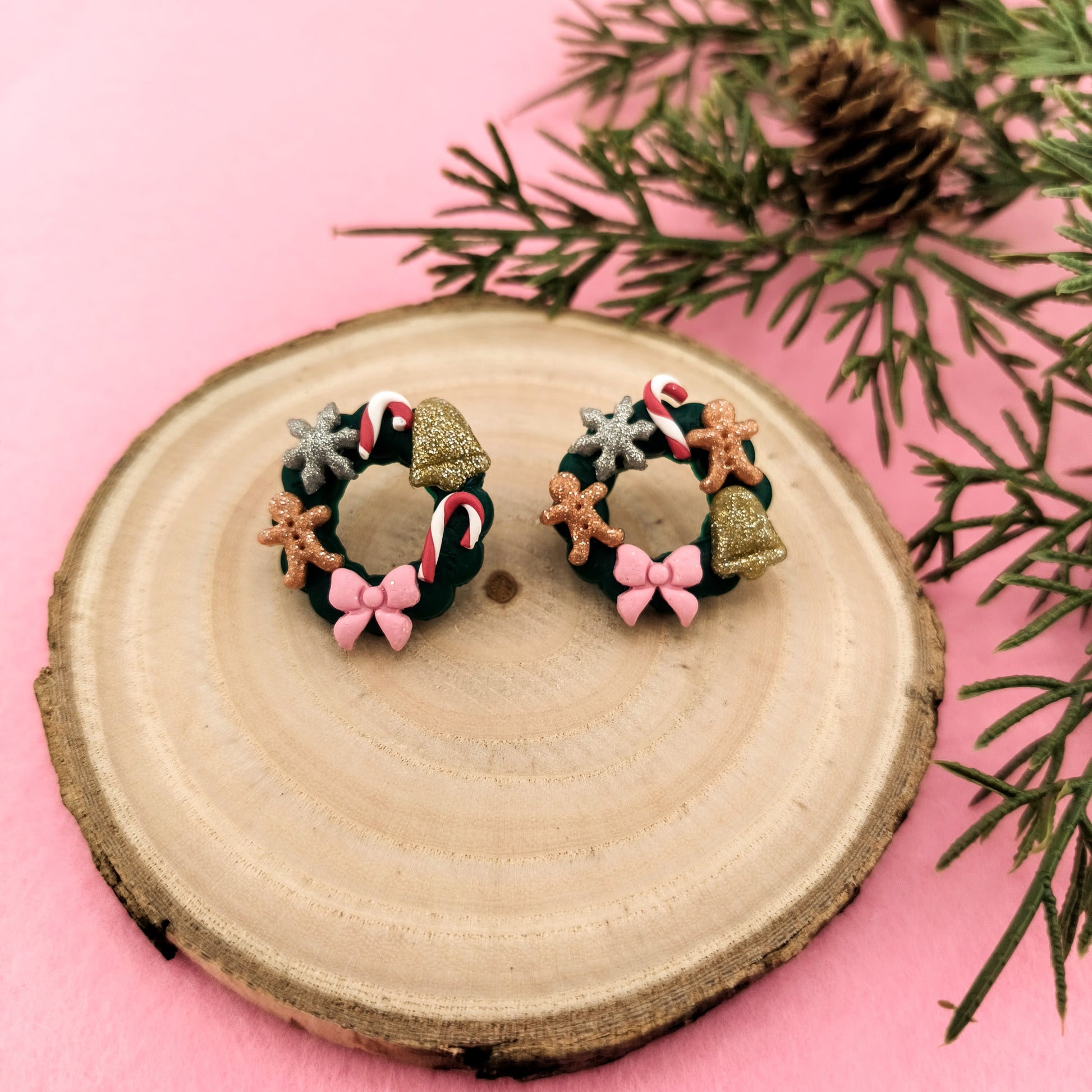 Pair of festive Christmas wreath earrings on a wooden slice with pink background and greenery