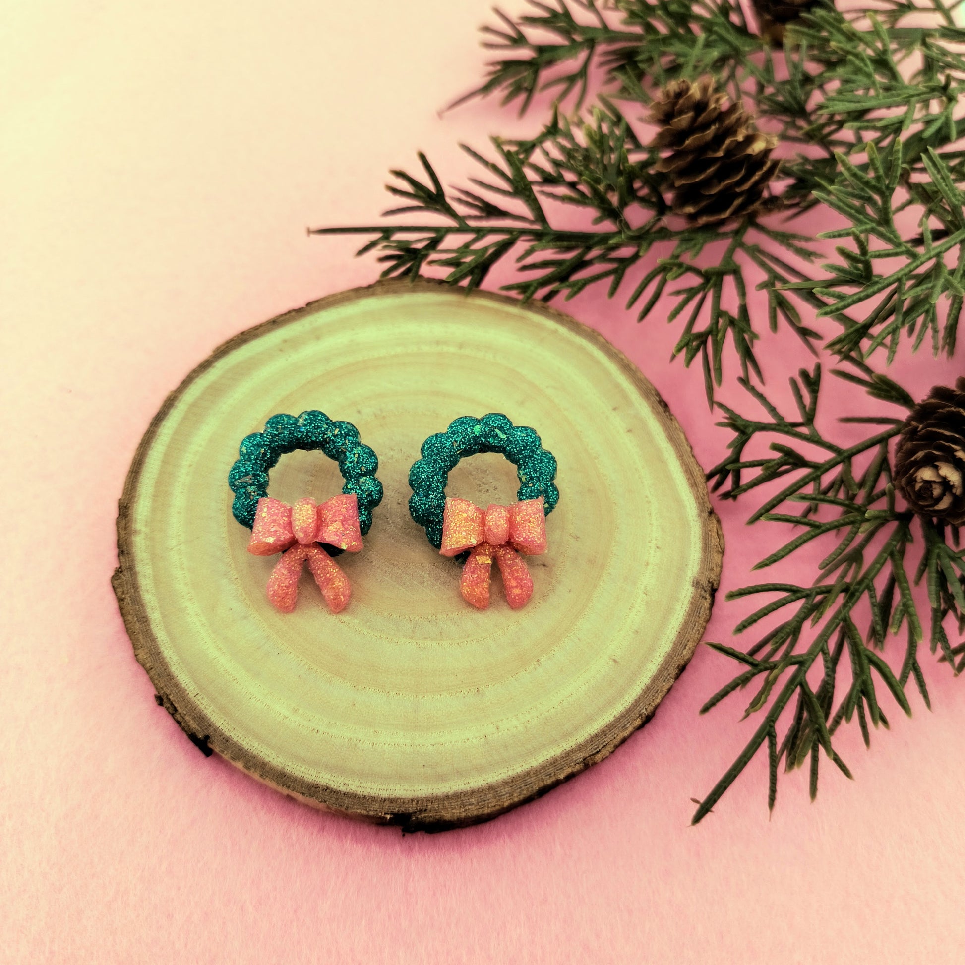 Pair of Christmas wreath earrings on a wooden slice with pine branches.
