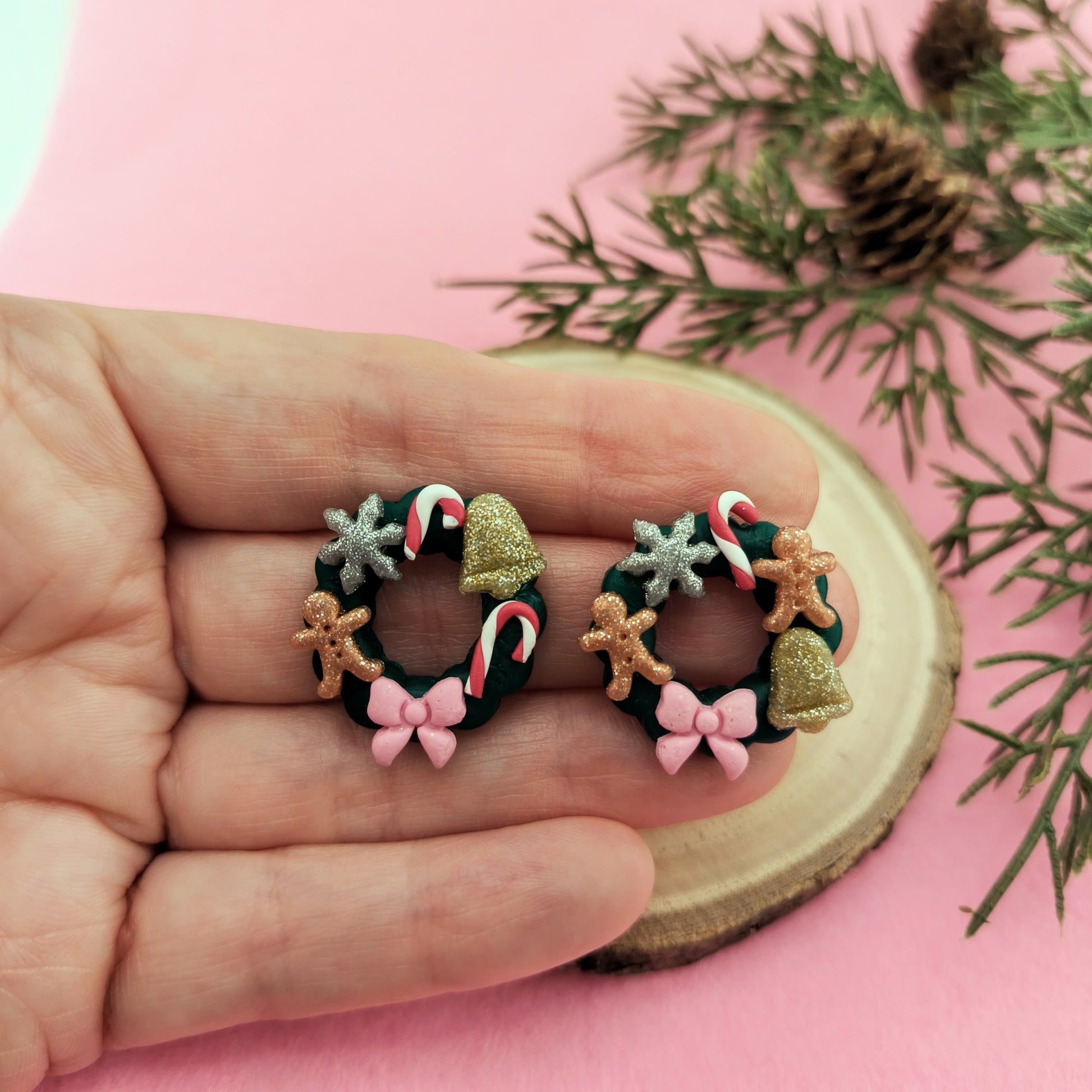 Small Christmas wreath earrings held in a hand with a pink background and greenery.