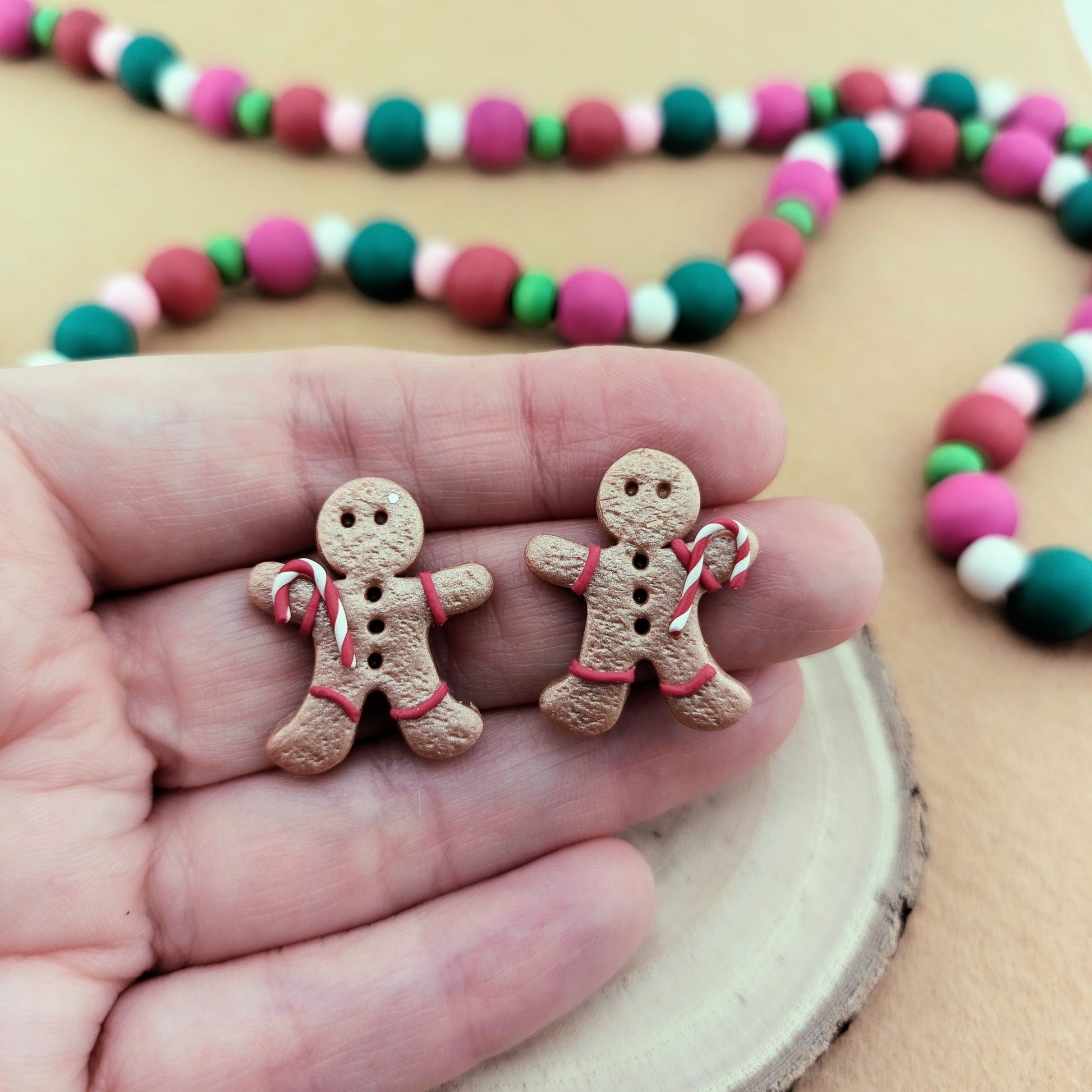 Two gingerbread man-shaped earrings held between fingers with a colorful beaded garland in the background.