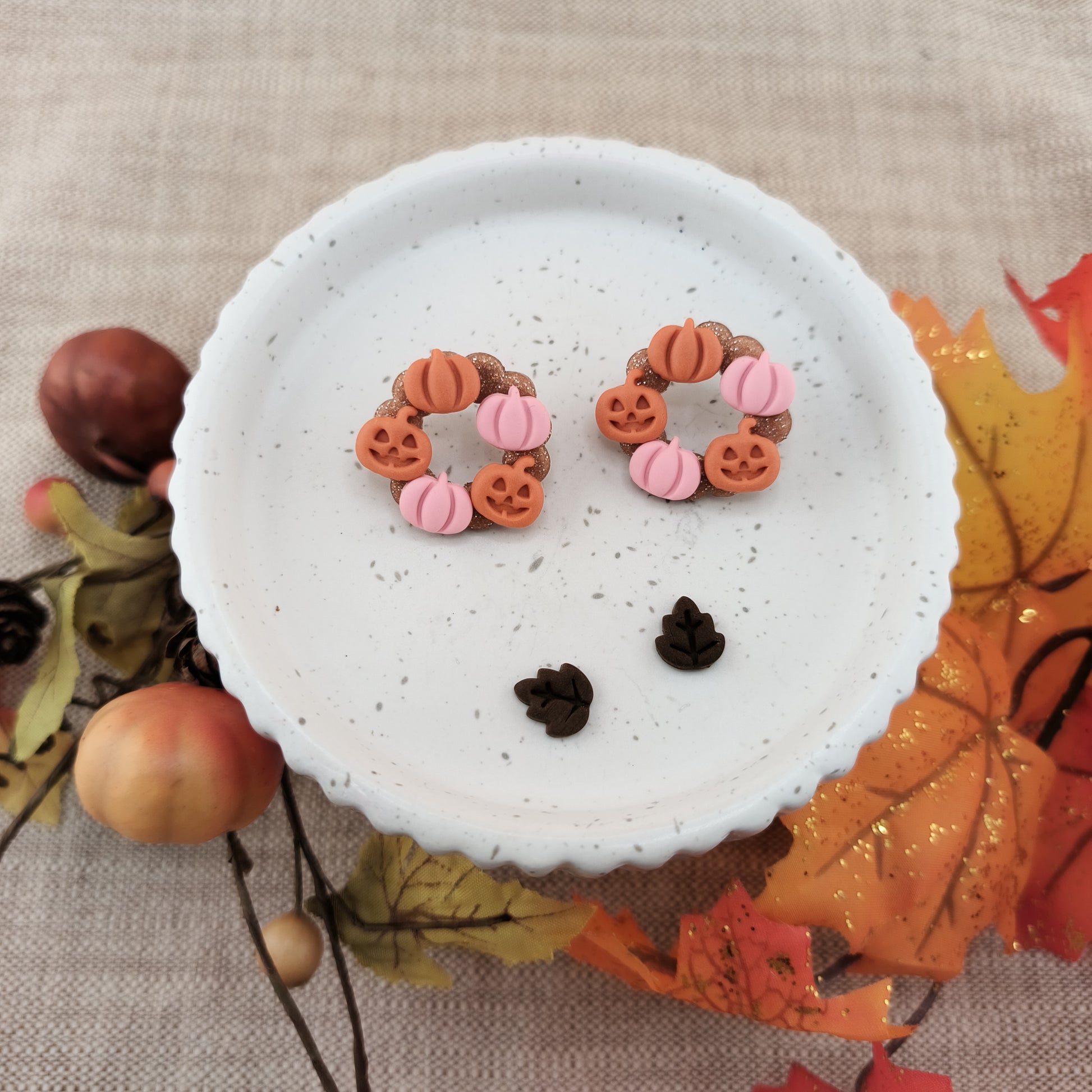 Pair of earrings with pumpkin and jack o lanterns on a white surface with autumn-themed decorations.