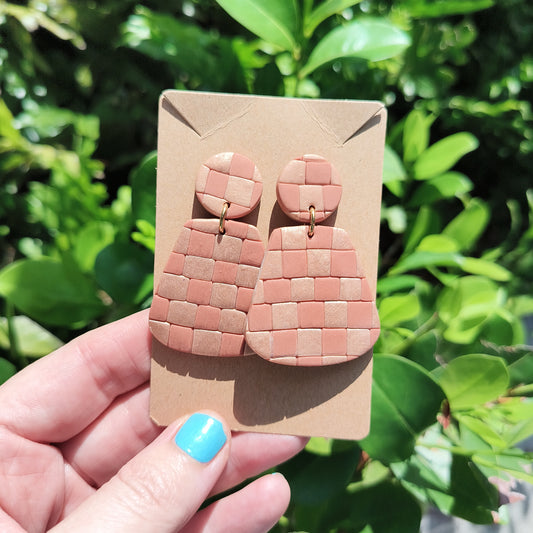 Checkerboard Rose Gold Polymer Clay Dangle Earrings Against green foliage.