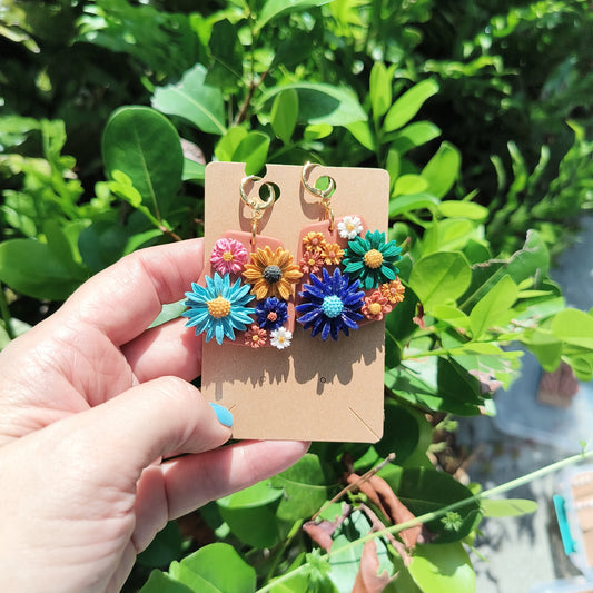 Polymer Clay dangle earrings covered in multicolored polymer clay flowers in different sizes, against a backdrop of green foliage.