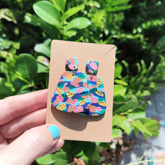 Multi colored, textured polymer clay dangle earrings against a backdrop of green foliage.