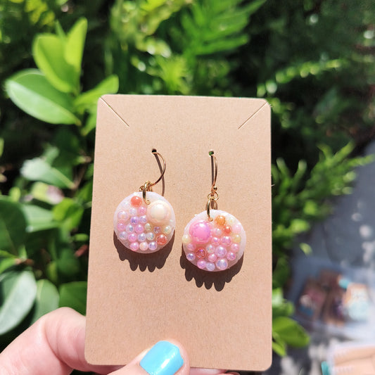 Cream and pink pearls cover small dangle earrings, against a backdrop of foliage.