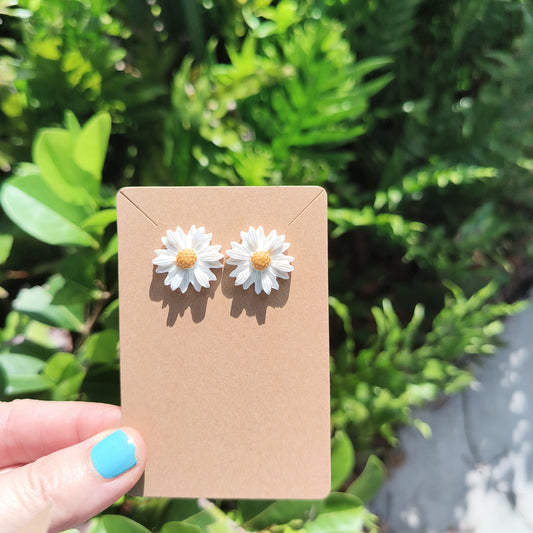 Medium sized white daisy polymer clay studs, against a backdrop of green foliage. 