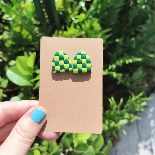 Green checkerboard polymer clay stud earrings, against a backdrop of green foliage.