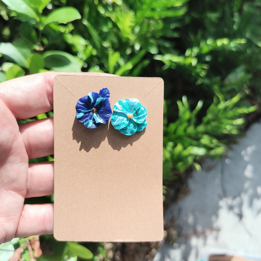 Mismatched blue clay flower stud earrings against a backdrop of green foliage.