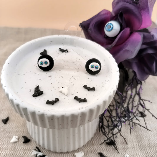 White pedestal with black decorations on a textured surface with purple flowers, displaying a pair of skull stud earrings.