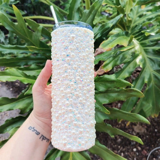 Stainless steel tumbler covered in pearl hearts, sparkly rhinestones, and pearls with clear lid and metal straw; against a backdrop of philodendron.