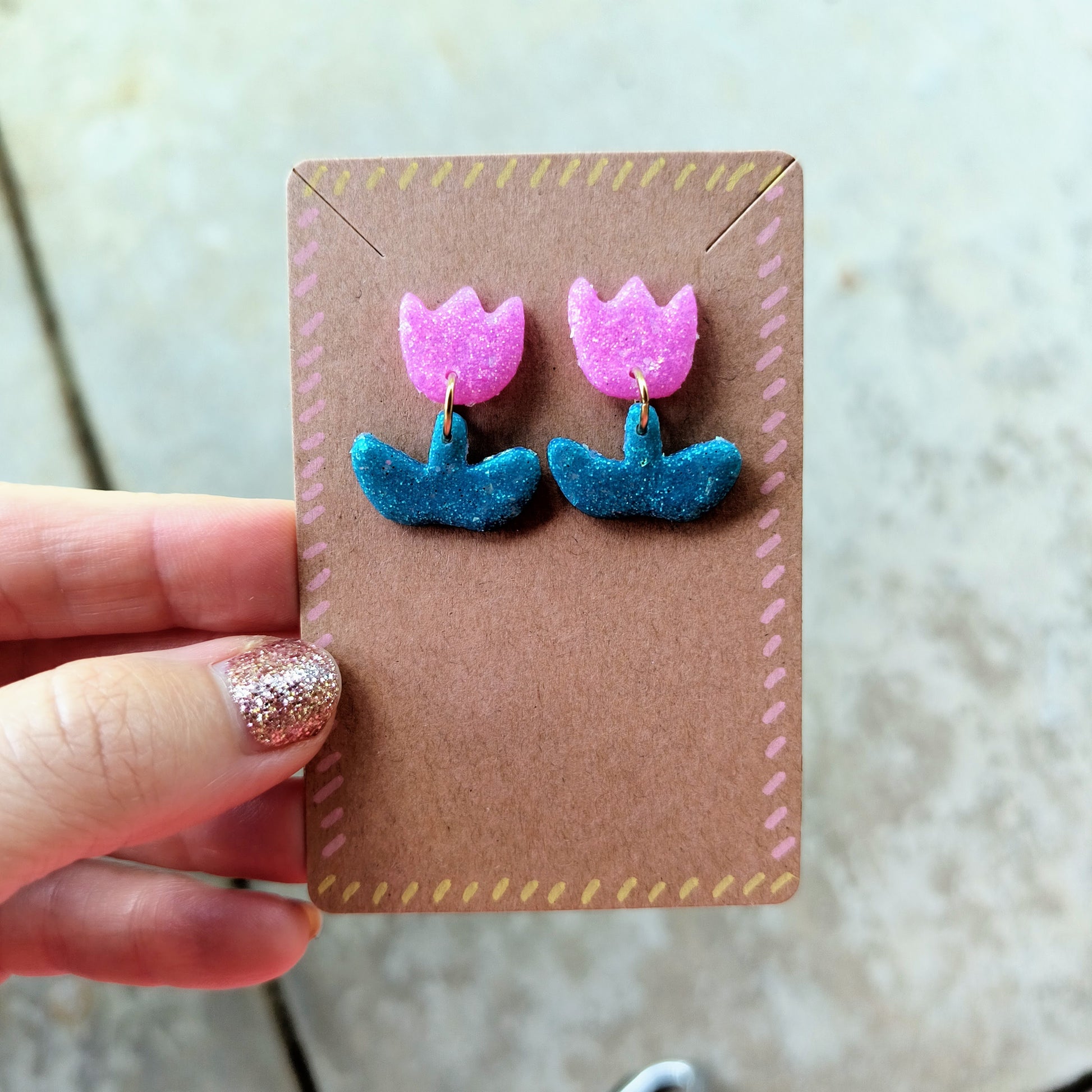 Pair of pink and green flower earrings on a brown card held by a hand.