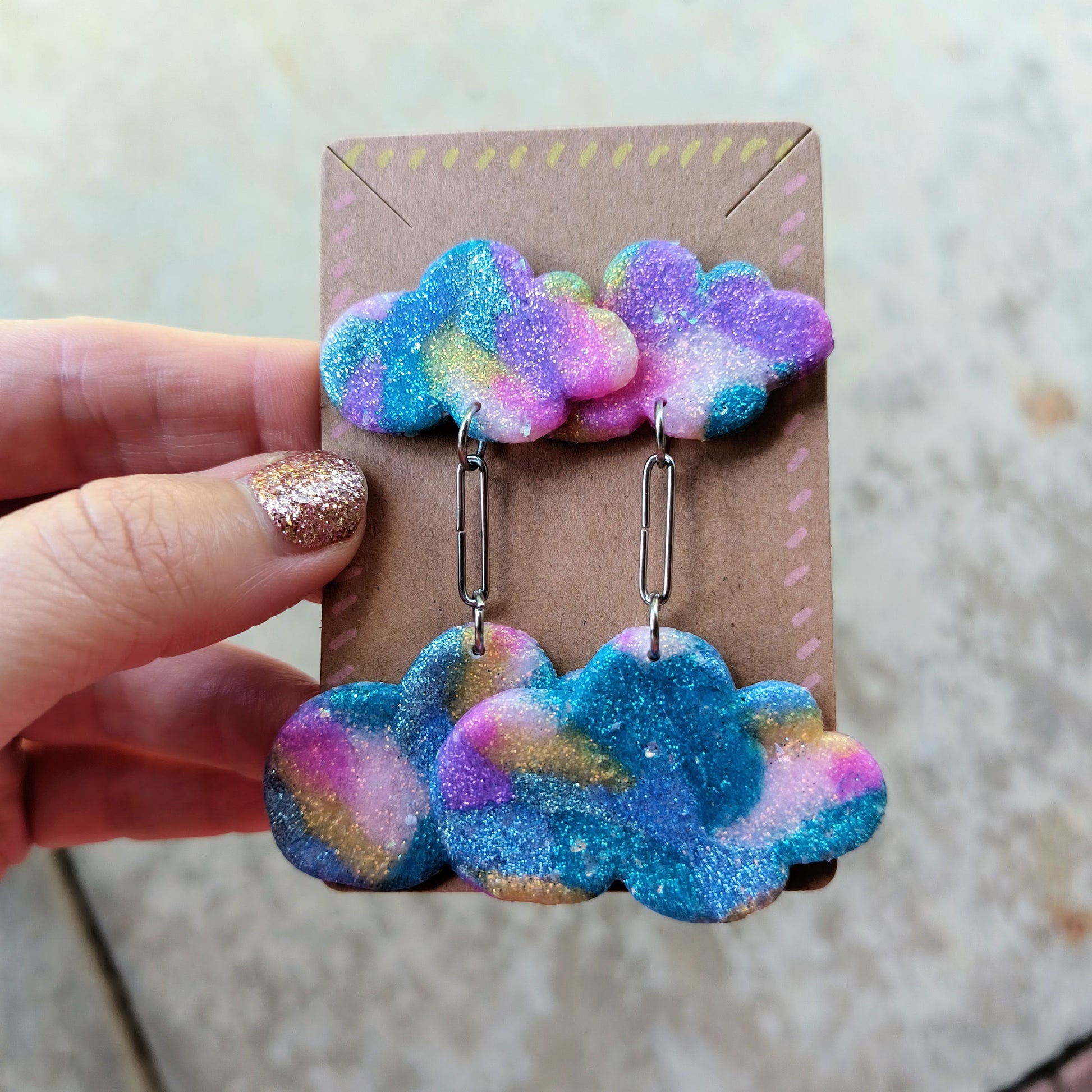 Colorful cloud-shaped earrings on a cardboard backing held by a hand.