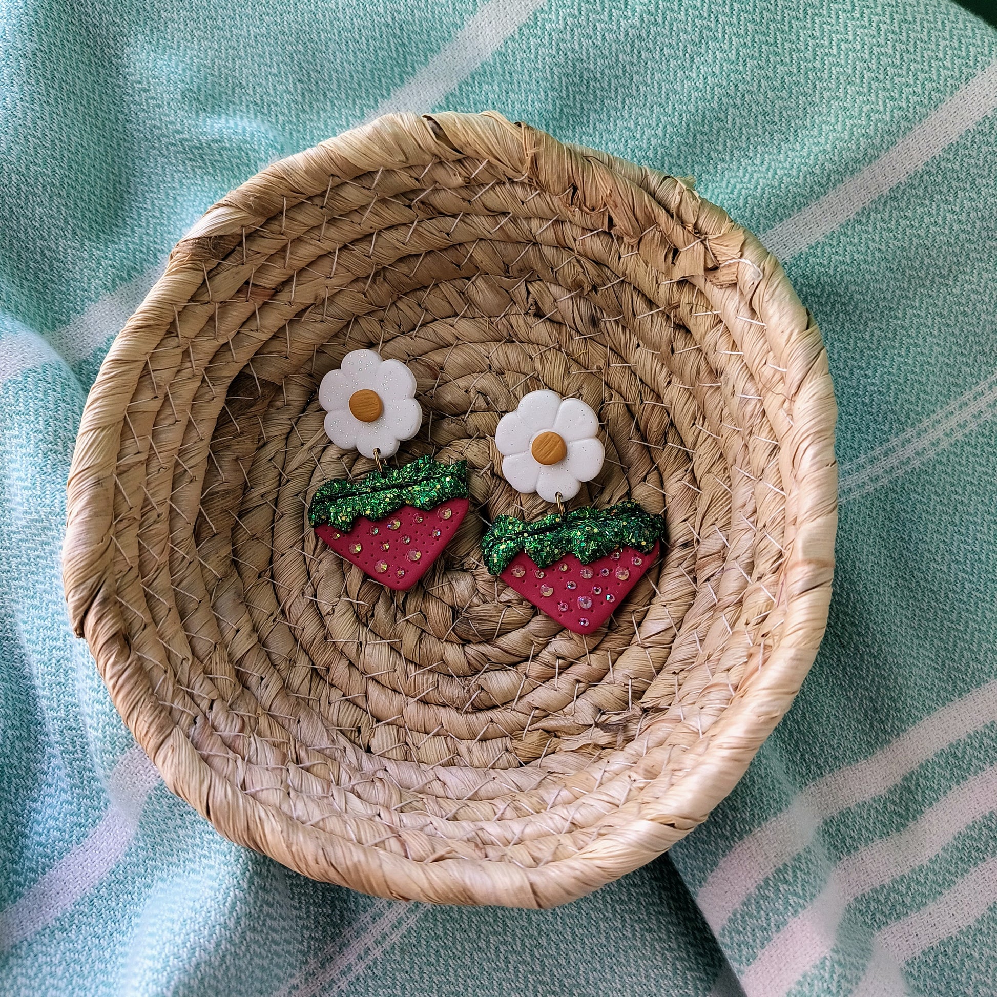 Woven dish with strawberry and flower earrings on a light blue fabric background