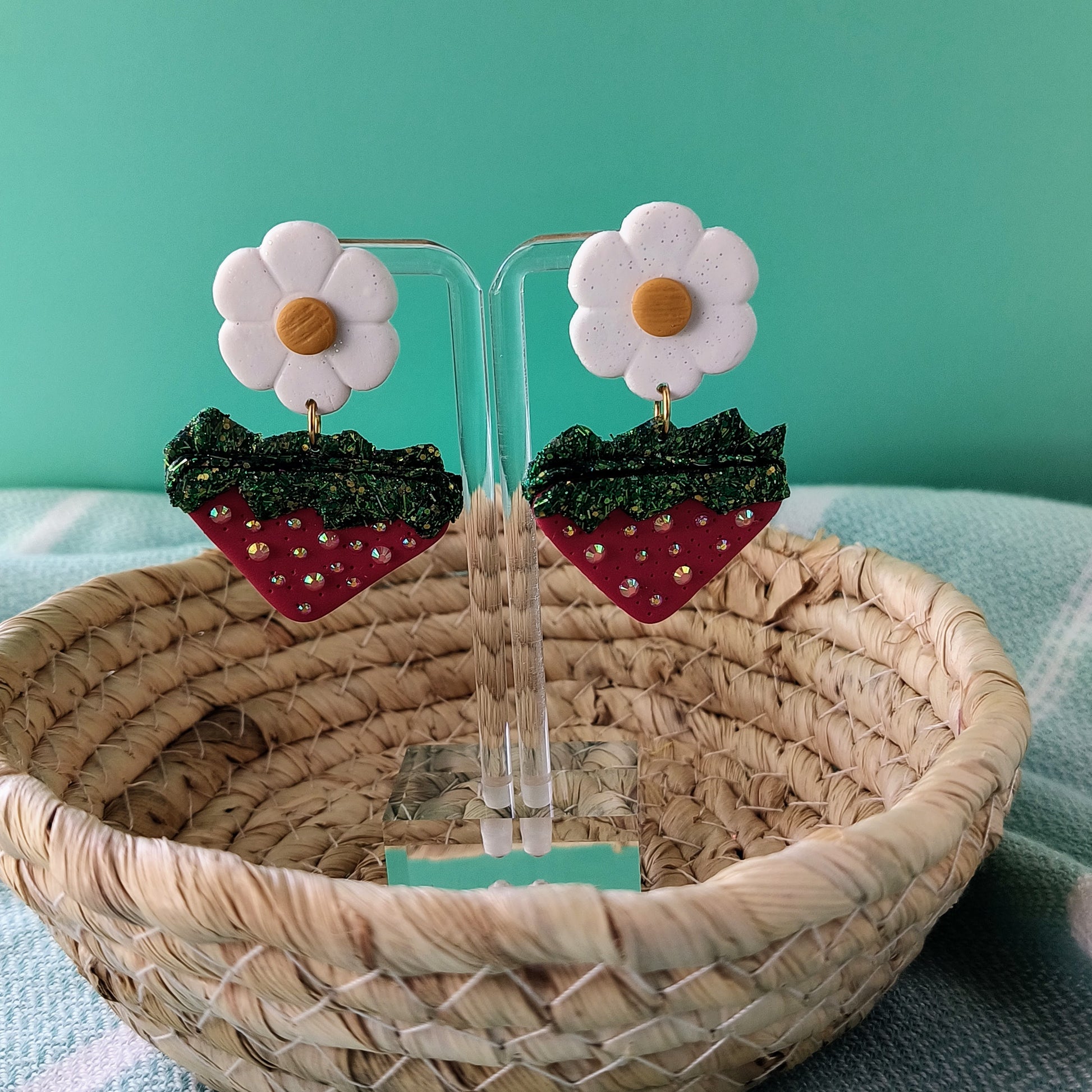 Strawberry and flower earrings on clear stands with a woven basket and teal background