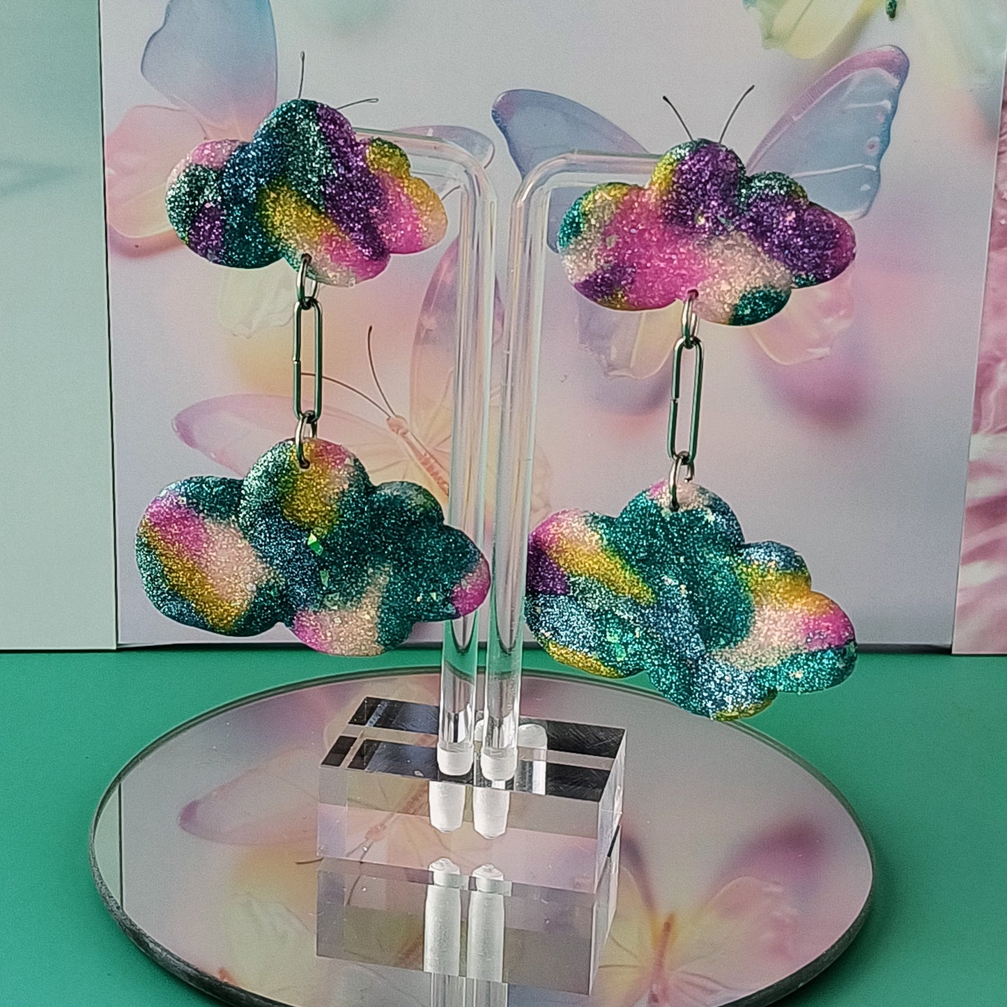 Colorful glittery cloud earrings on a clear stand with a soft, multicolored background.