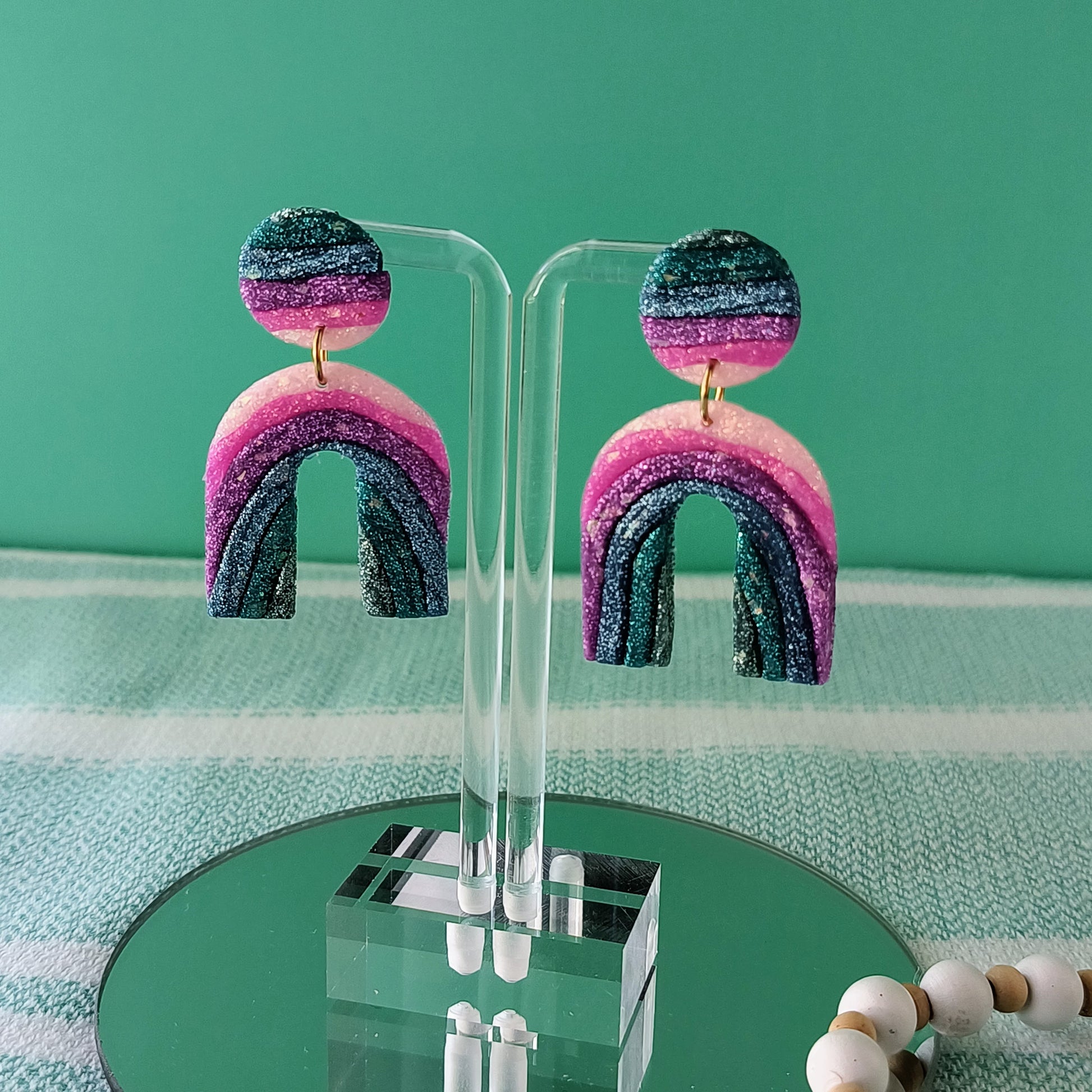Colorful sparkle arch-shaped earrings on a clear stand against a green background
