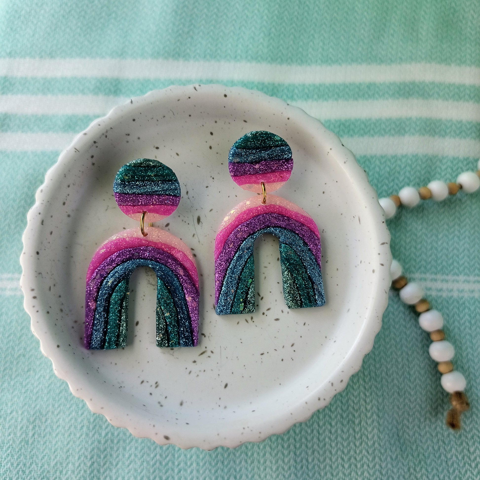 Pair of rainbow-shaped sparkle earrings on a white dish with a light blue background