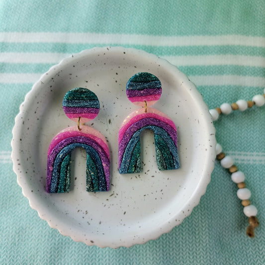 Pair of rainbow-shaped sparkle earrings on a white dish with a light blue background