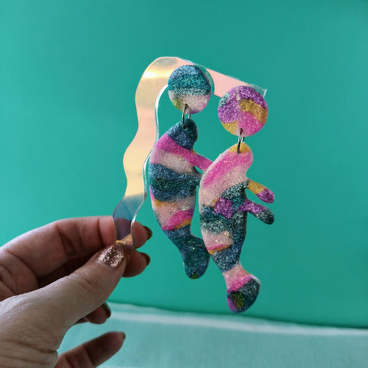 Colorful glittery manatee earrings held by a hand against a teal background