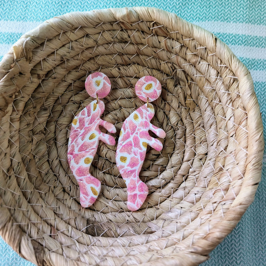 Pink and white patterned manatee earrings on a woven mat with a blue and white striped background